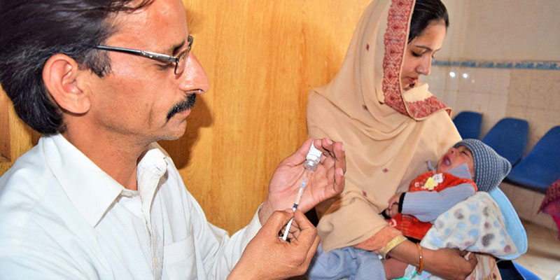 Farwa holding her baby while the vaccinator prepares an injectable polio vaccine